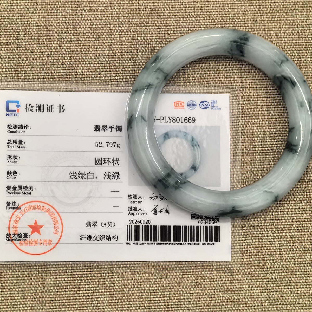 White and dark green jadeite bangle displayed with its matching NGTC certificate on a woven background.