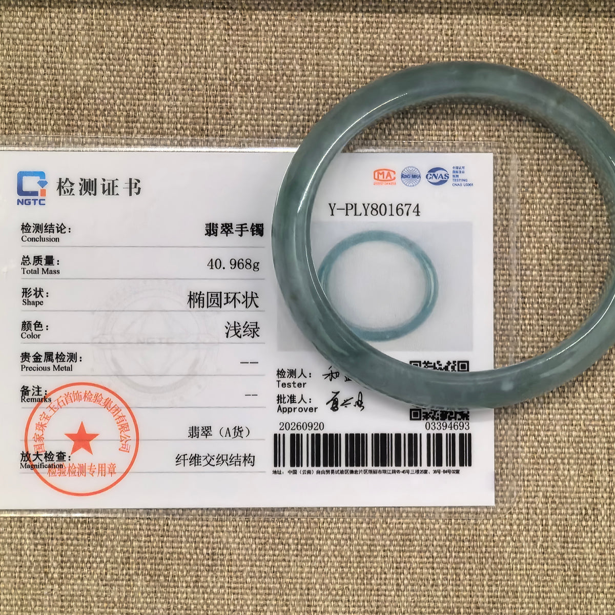 Blue-green oval jadeite bangle displayed with its matching NGTC certificate on a woven background.
