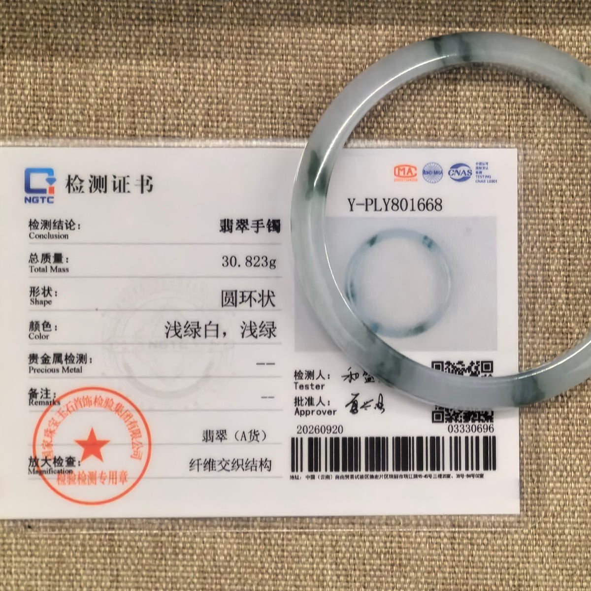 Milky white jadeite bangle with green floating patterns displayed with its matching NGTC certificate on a woven background.