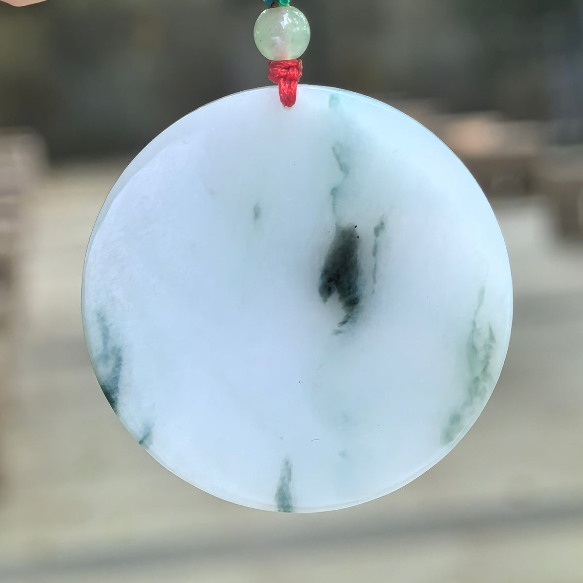 White jadeite round plain plaque pendant suspended from a cord, with a deep green central patch and finer green wisps across its uncarved face.