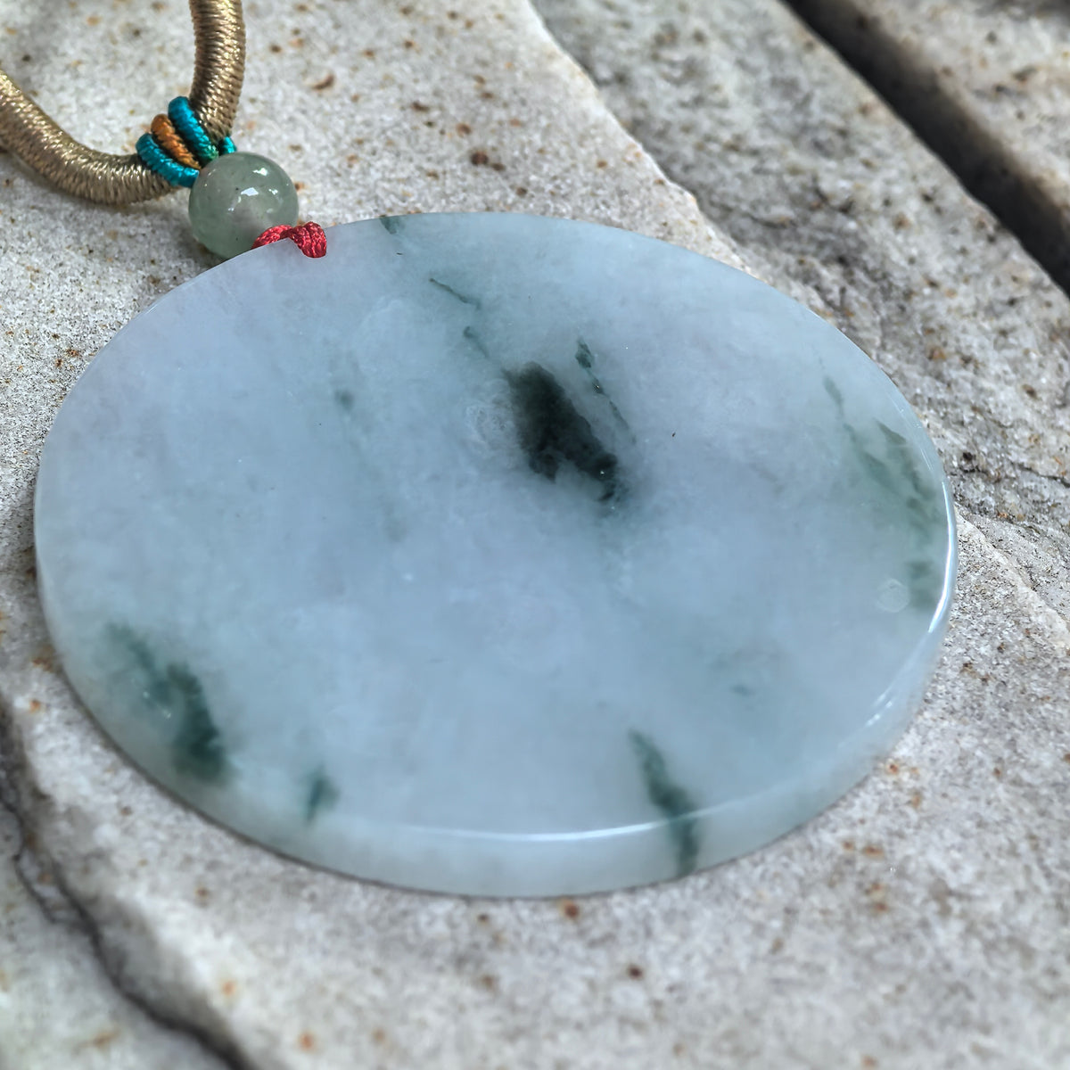 Angled view of a white jadeite round plain plaque pendant resting on pale stone, showing deep green floating patterns and a polished edge.