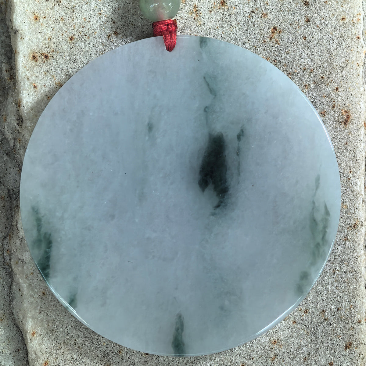 Top view of a fine glutinous jadeite round plain plaque pendant on stone, showing a white base with scattered deep green patterns.