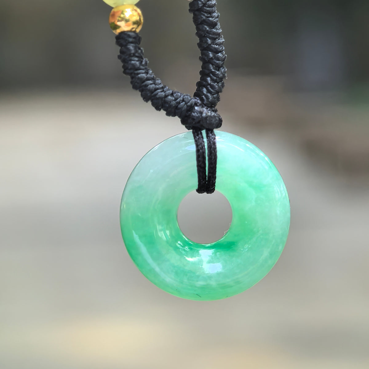 Close-up of a jadeite donut pendant suspended from a black cord, showing pale green areas, brighter green accents, and a polished circular opening.