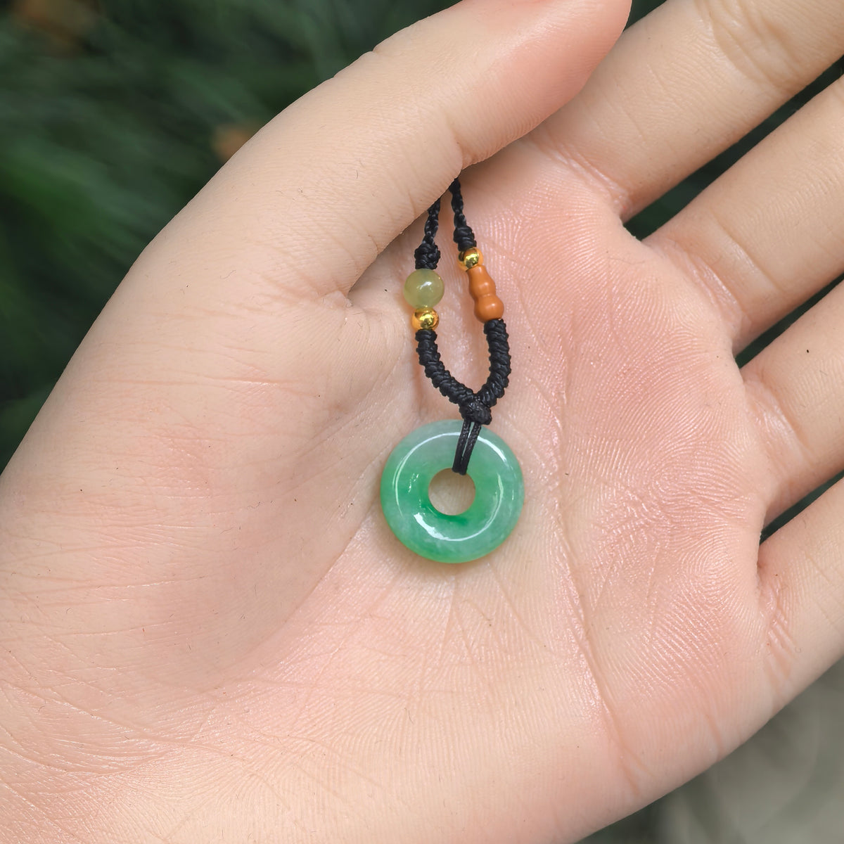 Petite white and green jadeite donut pendant resting in an open palm, attached to a black knotted cord with decorative beads.