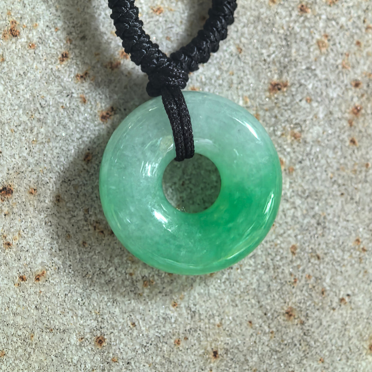 Top view of a white and green jadeite donut pendant resting on stone, with a black cord threaded through its central opening.