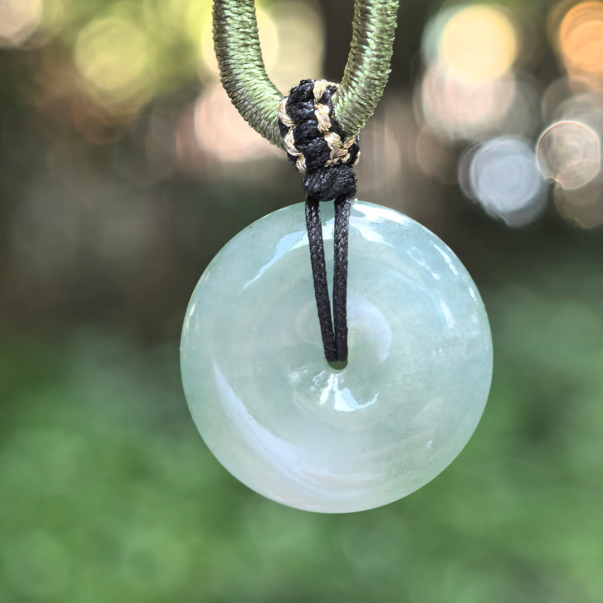 Jadeite peace pendant suspended from a decorative cord, showing a pale green glow, translucent texture, and polished circular surface.