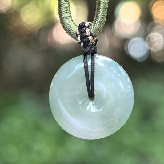 Jadeite peace pendant suspended from a decorative cord, showing a pale green glow, translucent texture, and polished circular surface.