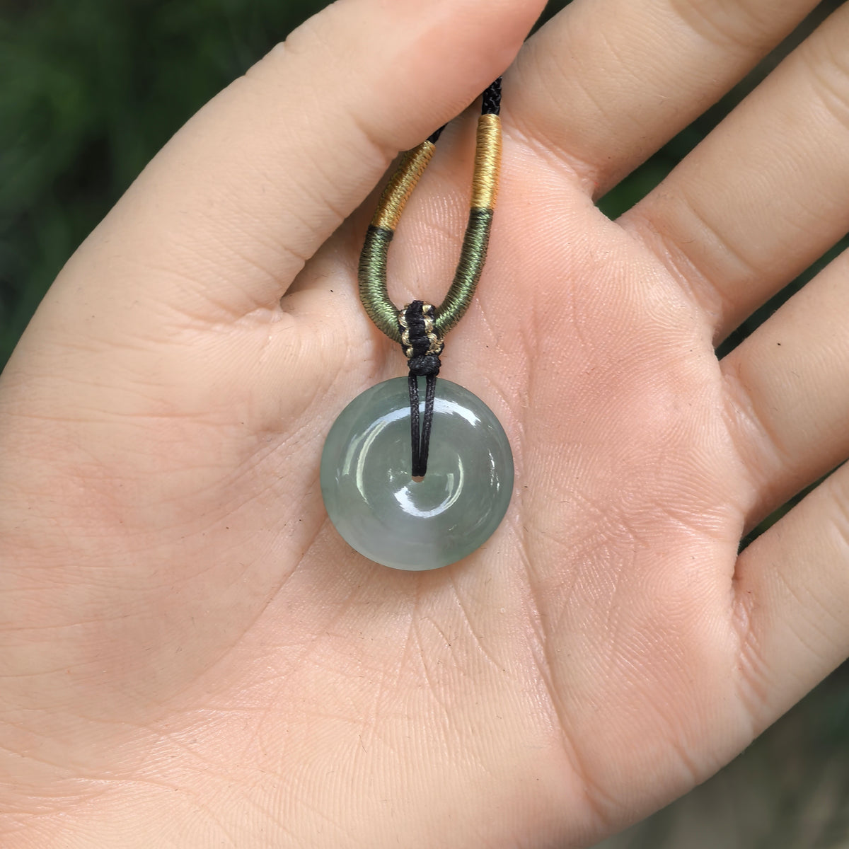 Gray-green jadeite peace pendant resting in an open palm, showing its proportions and cord wrapped with green and gold-colored thread.