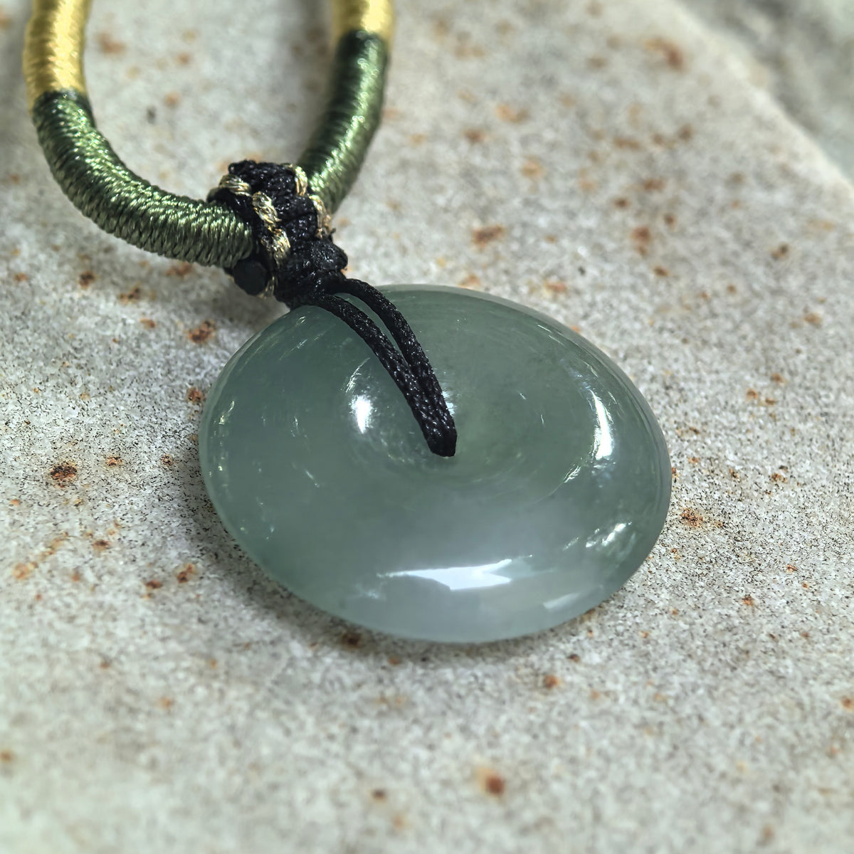Angled view of a gray-green jadeite peace pendant resting on pale stone, showing its curved profile and black cord attachment.