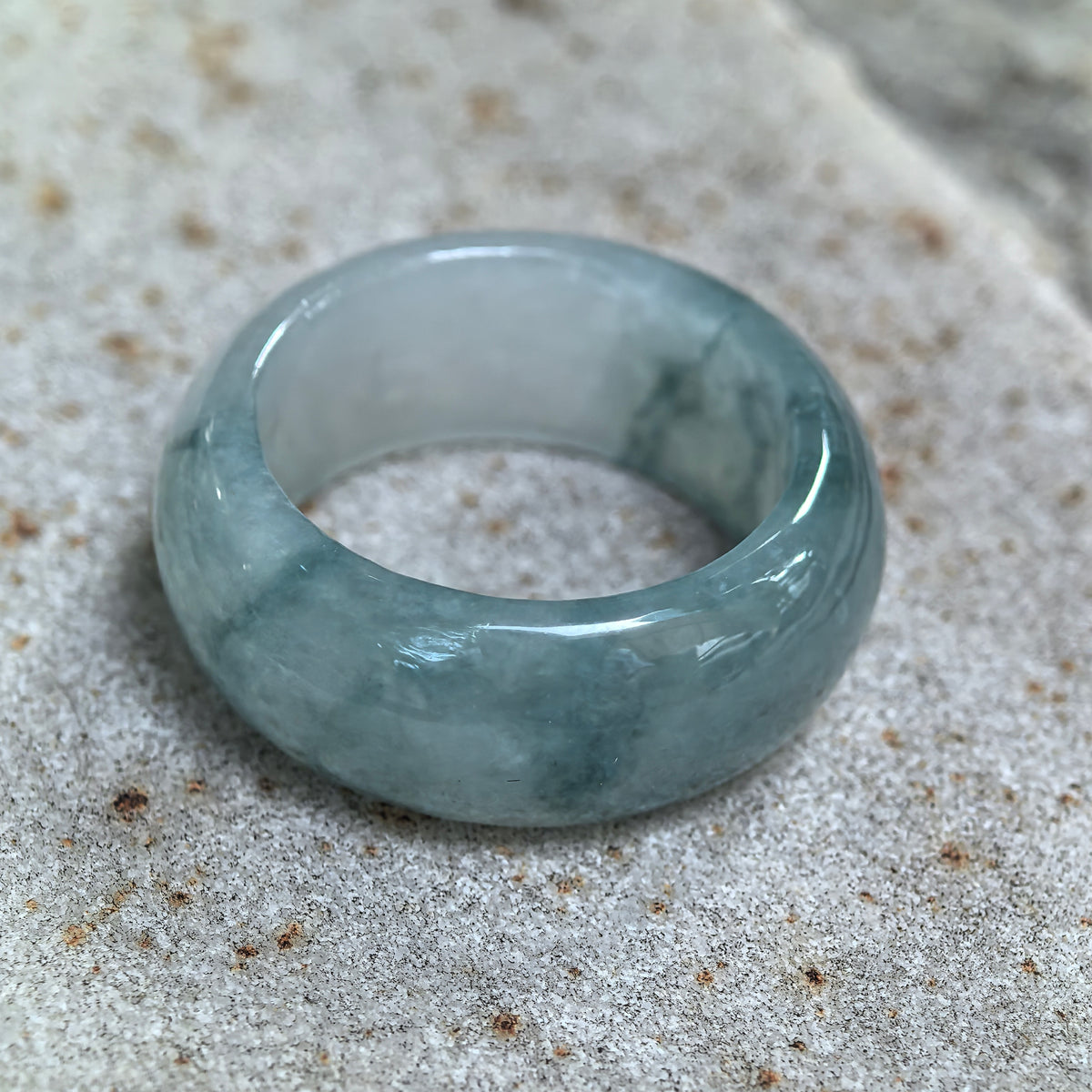 Wide jadeite ring band resting at an angle on pale stone, showing green floating patterns, a rounded exterior, and the inner surface.