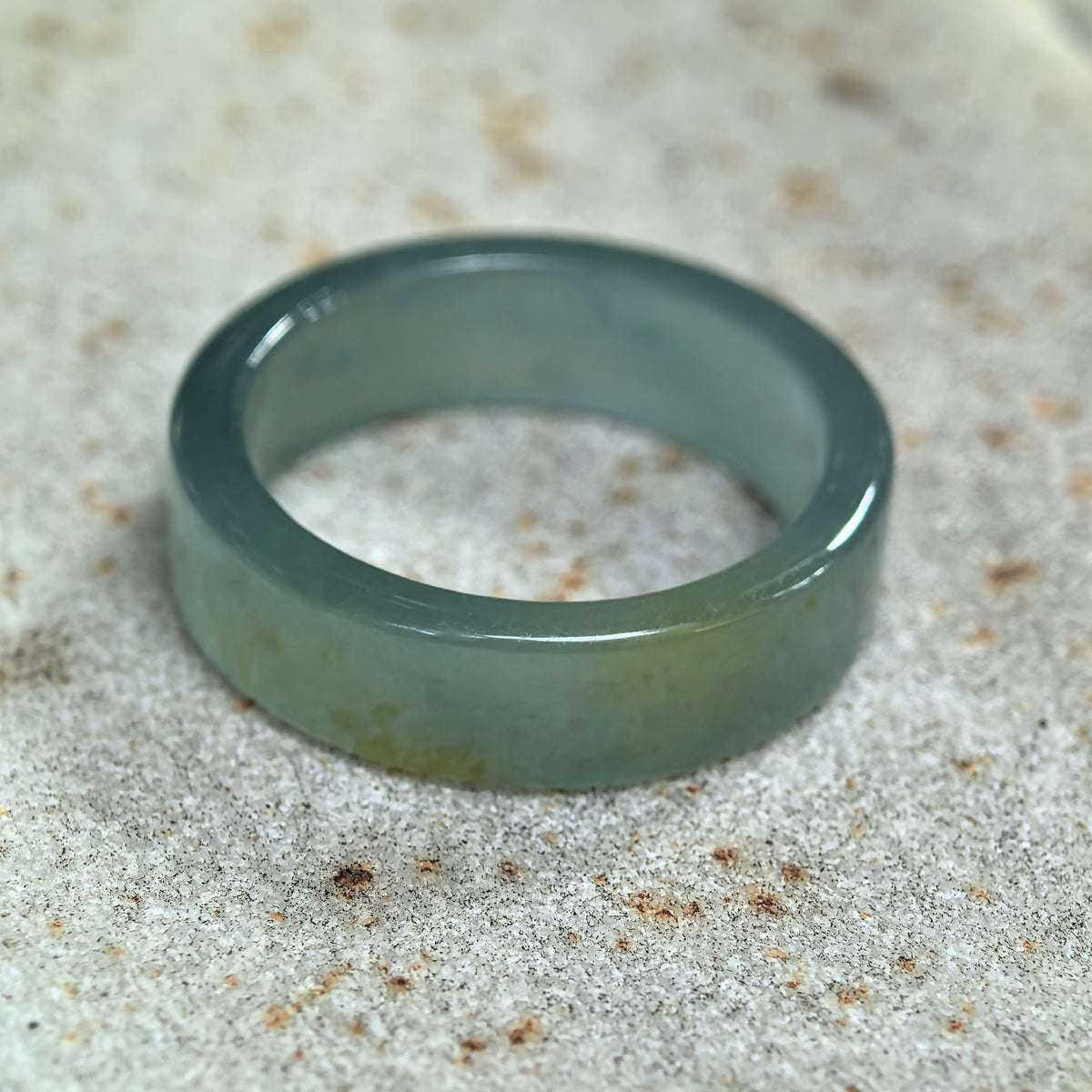 Green jadeite ring band resting at an angle on pale stone, showing its straight-cut profile, inner surface, and yellow accent.