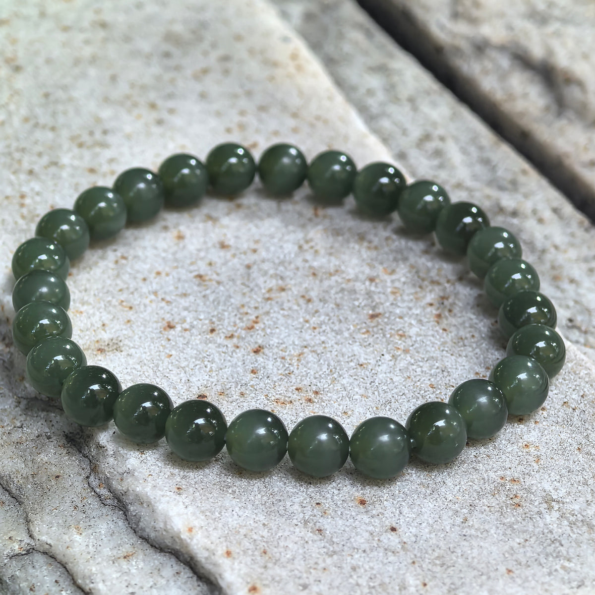 Deep green Hetian jade bead bracelet resting on pale stone, viewed at an angle to show the rounded beads and polished luster.