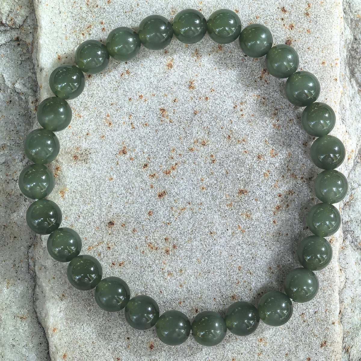 Top view of a dark green nephrite bead bracelet arranged in a circle on stone, showing subtle lighter bands across some beads.