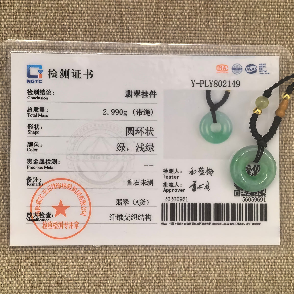 Green jadeite donut pendant and black cord displayed with the matching NGTC certificate on a woven background.