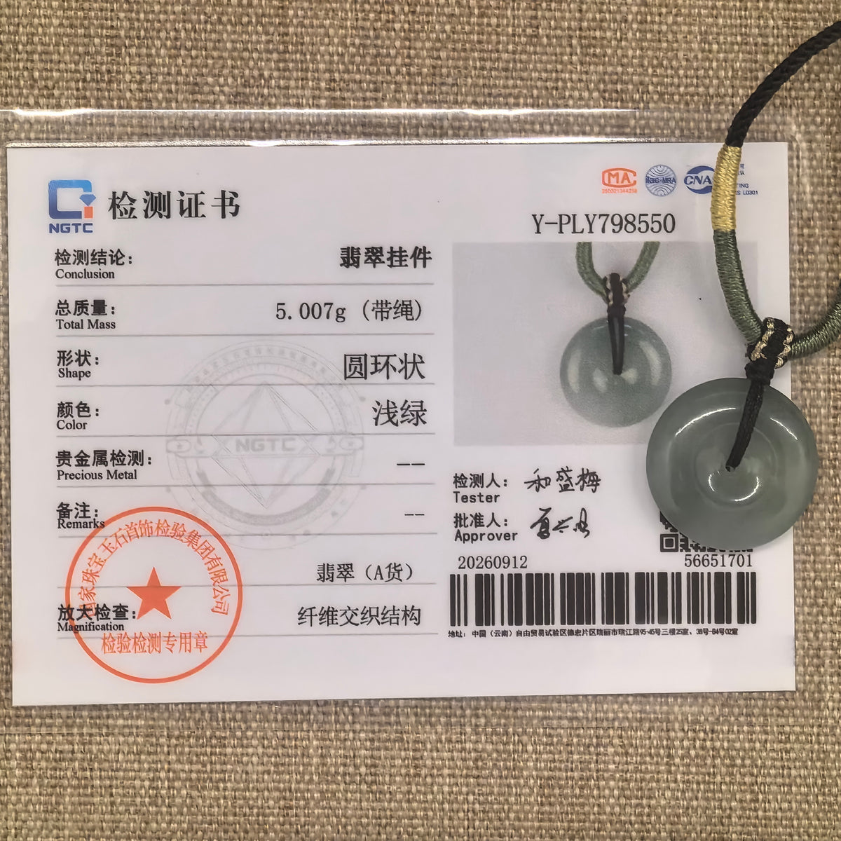 Gray-green jadeite peace pendant displayed with its matching NGTC certificate on a woven background.