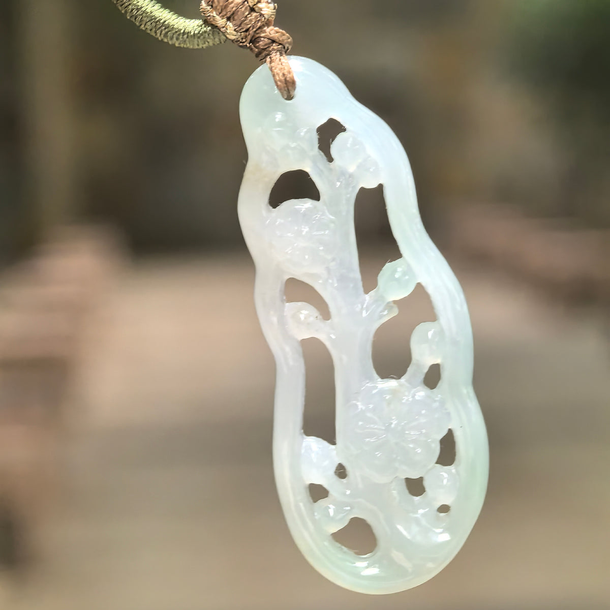 Suspended pale green jadeite plum blossom pendant in natural light, highlighting its icy glutinous translucency and carved flowers and buds.