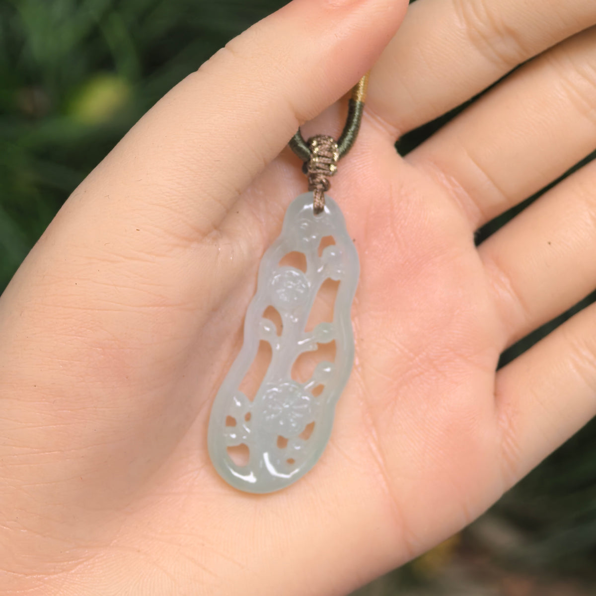 Pale green jadeite plum blossom pendant resting on an open palm, showing its elongated openwork design and decorative knotted cord.