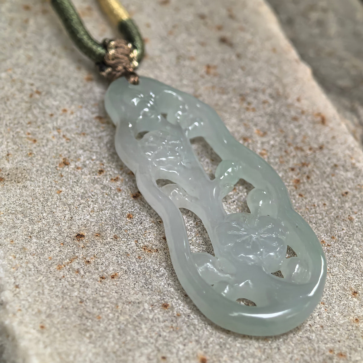 Angled close-up of a pale green jadeite plum blossom pendant on stone, showing the carved petals, openwork branches, and polished edge.