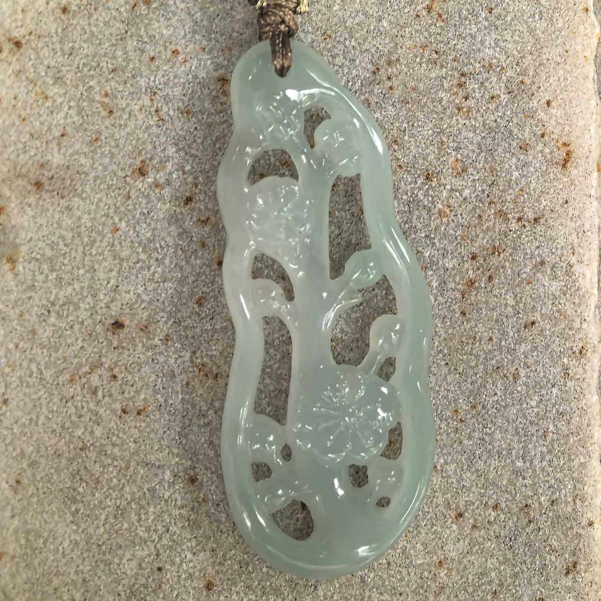 Front view of a pale green jadeite pendant on a stone surface, showing plum blossoms and branching stems framed by openwork cutouts.