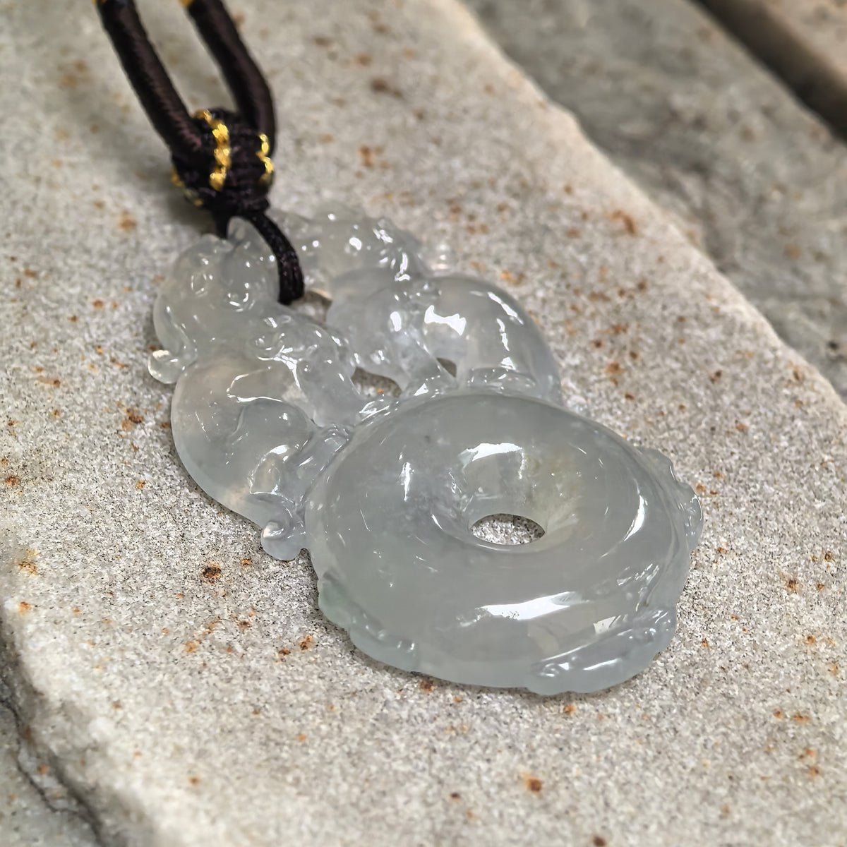 Angled close-up of a pale green jadeite double Pixiu pendant on stone, showing the raised carving and rounded peace pendant base.