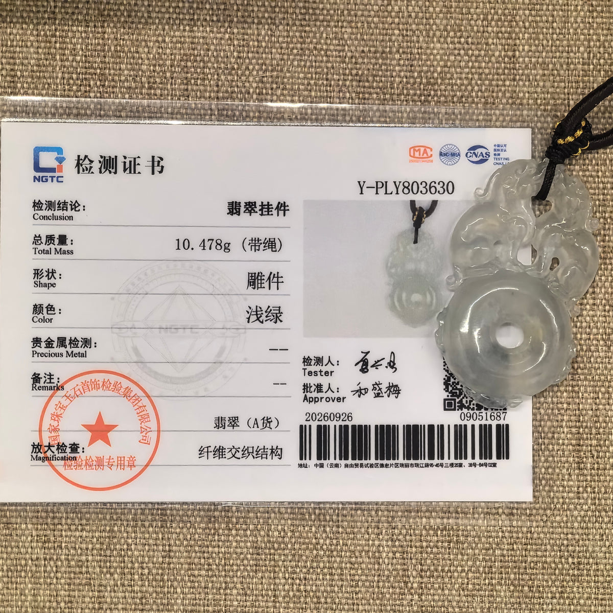 Double Pixiu jadeite peace pendant displayed beside its matching NGTC certificate on a woven background.