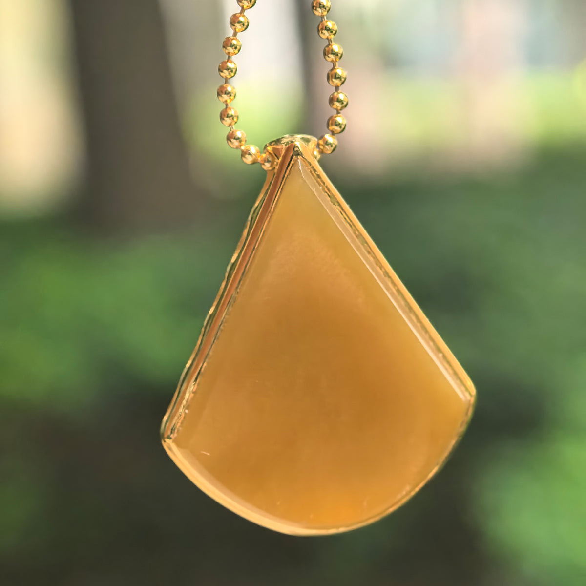 Yellow jadeite teardrop pendant in an 18K gold setting, suspended from a gold-tone beaded chain against a blurred green background.