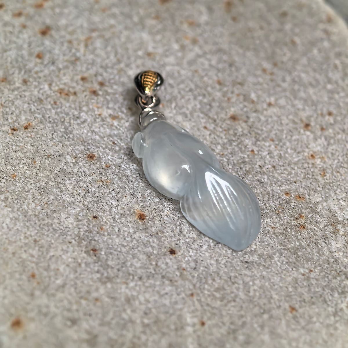 Angled close-up of a white jadeite goldfish pendant on stone, highlighting its rounded body and curved tail grooves.