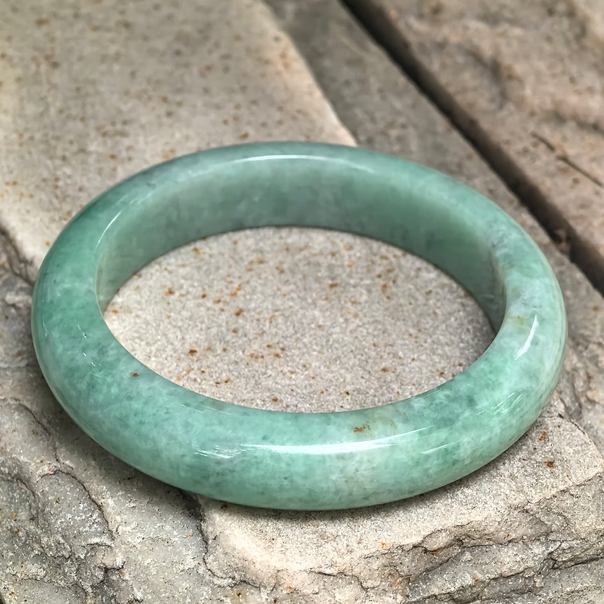 Angled view of an apple green D-shape jadeite bangle on stone, showing its flatter inner profile and small brown flecks.