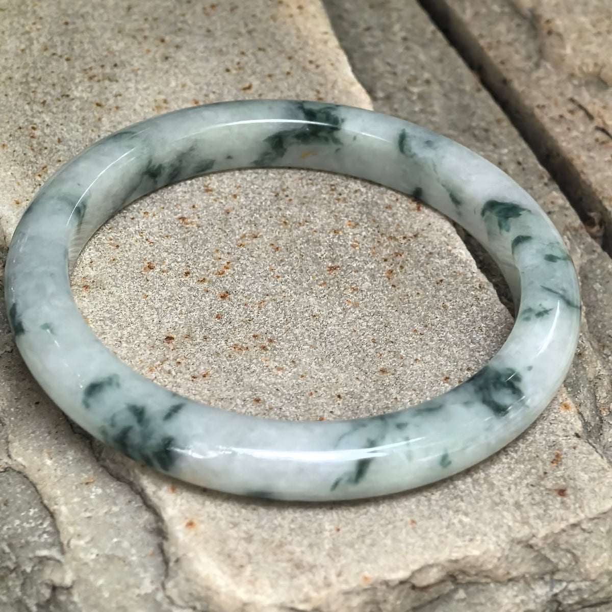 Angled view of a slim D-shape jadeite bangle on stone, showing its flatter inner profile and small golden-brown flecks.