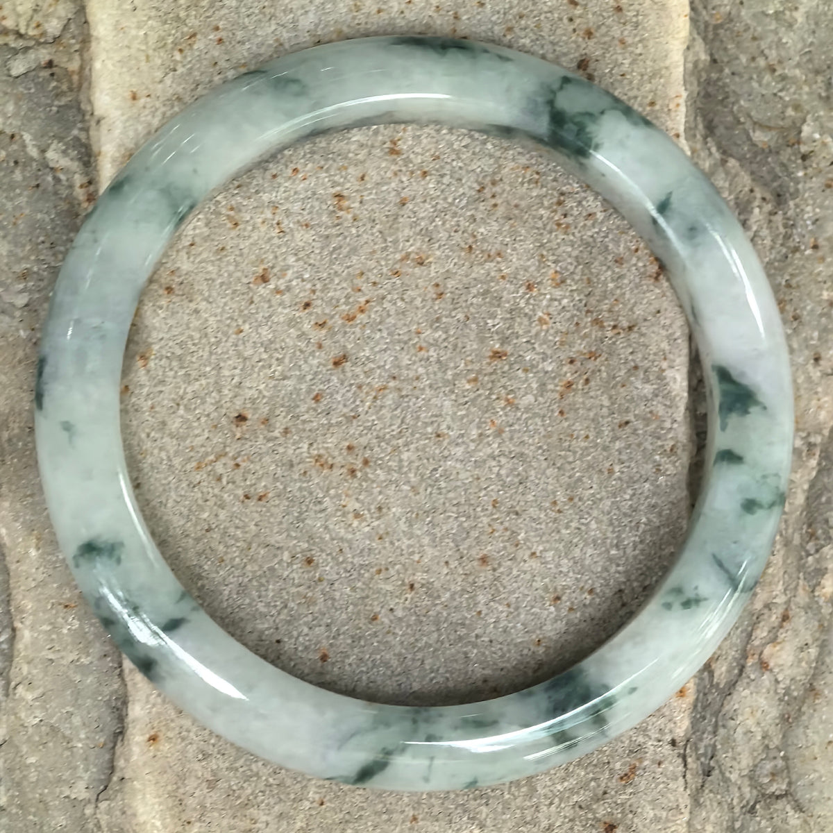 Overhead view of a milky white jadeite bangle on stone, showing deep green floating patterns around the circular band.
