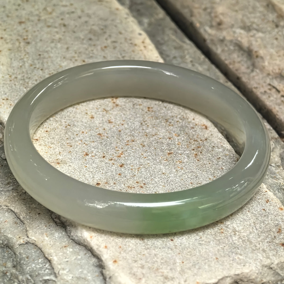 Angled view of a greenish white nephrite D-shape bangle resting on stone, showing its flat inner surface, rounded exterior, and green accent.