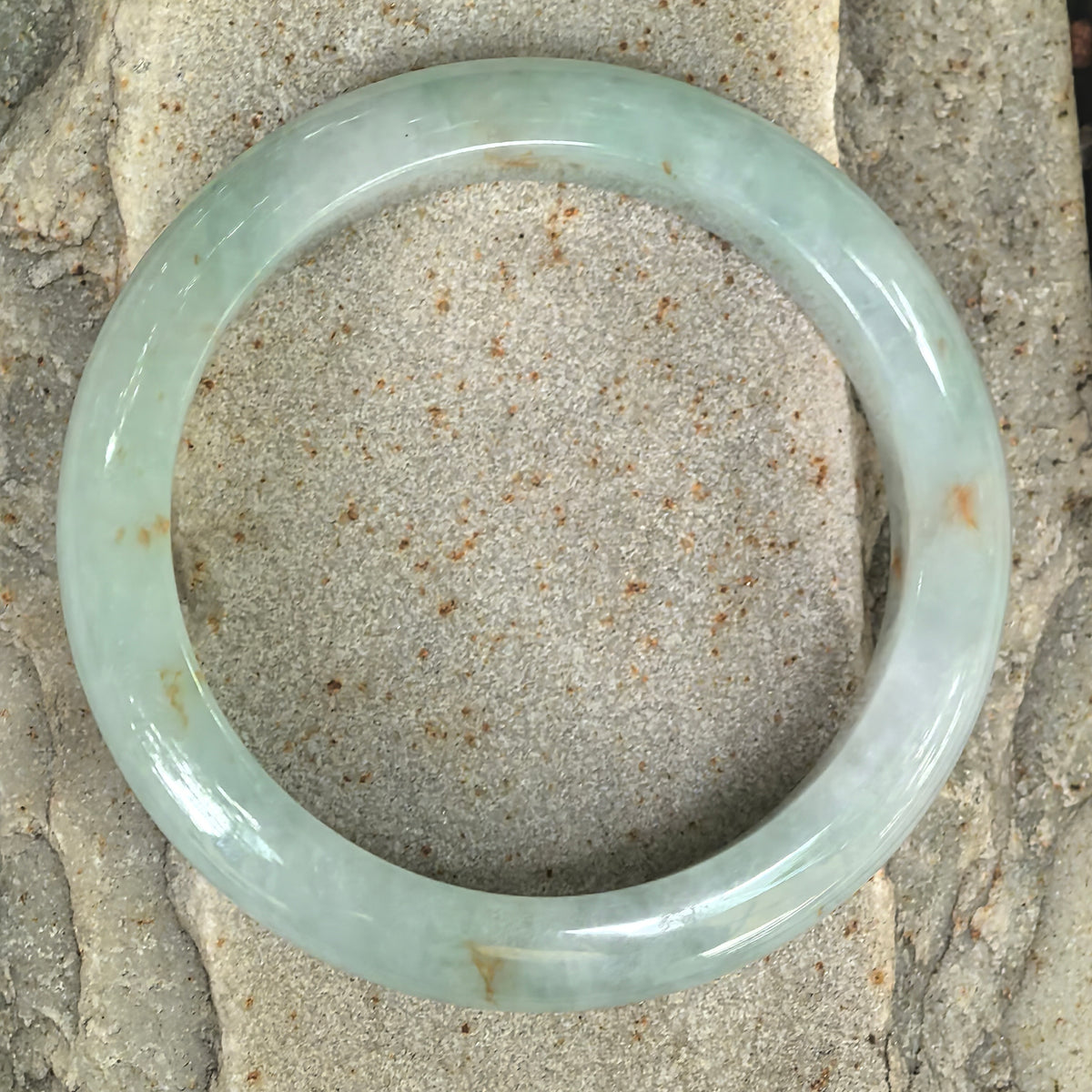 Overhead view of a pale apple green jadeite bangle on textured stone, showing its full circular shape and golden-brown flecks.