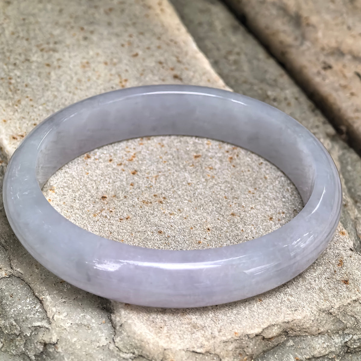 Angled view of a pale lavender oval jadeite bangle on textured stone, showing its rounded exterior and natural texture.