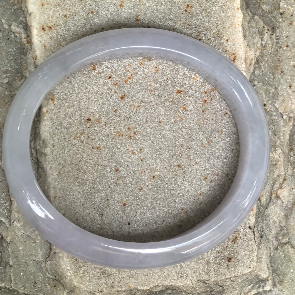 Overhead view of a pale lavender jadeite bangle on stone, showing its oval outline and polished surface.