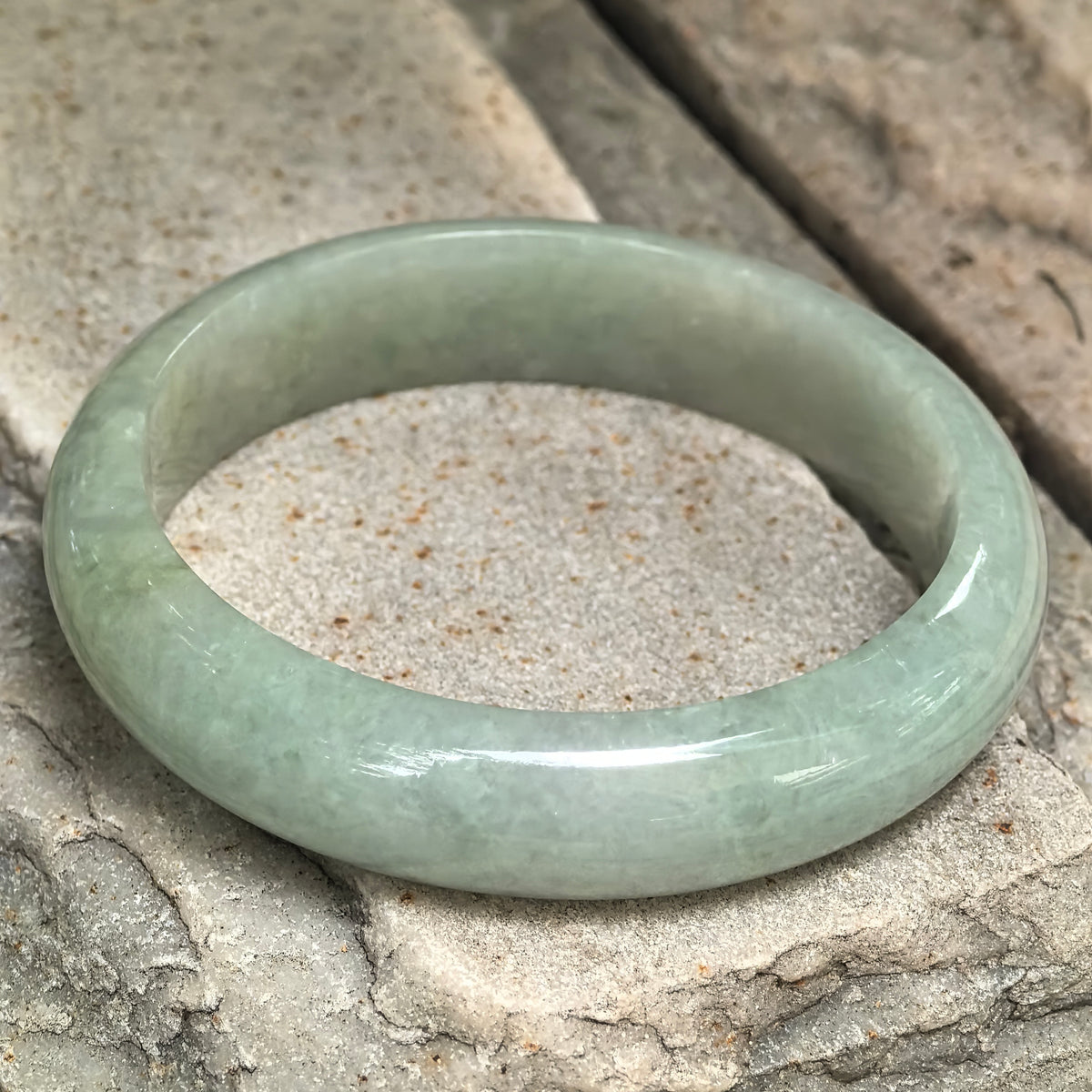 Angled view of a pale apple green jadeite D-shape bangle on textured stone, showing its rounded exterior and flatter inner profile.