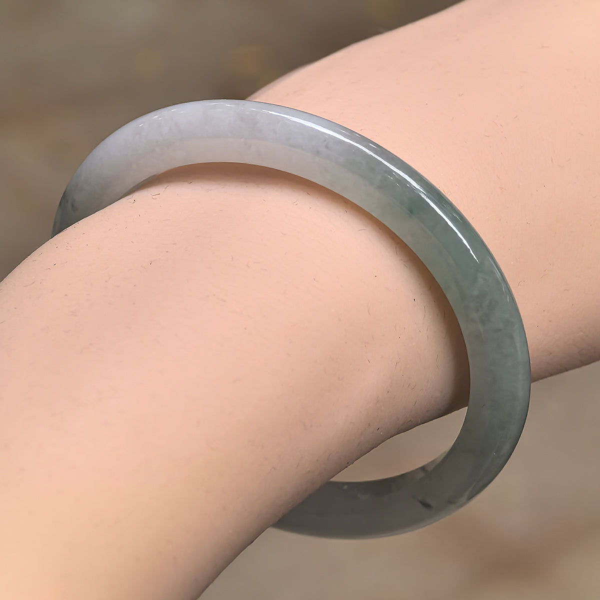Angled wrist view of a slim oval jadeite bangle, showing a pale section transitioning into green and a glossy polished surface.