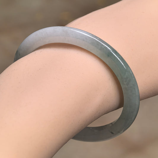 Angled wrist view of a slim oval jadeite bangle, showing a pale section transitioning into green and a glossy polished surface.