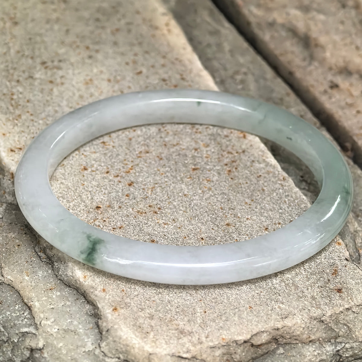 Slim oval jadeite bangle resting at an angle on textured stone, showing contrasting pale and green sections and smooth contours.