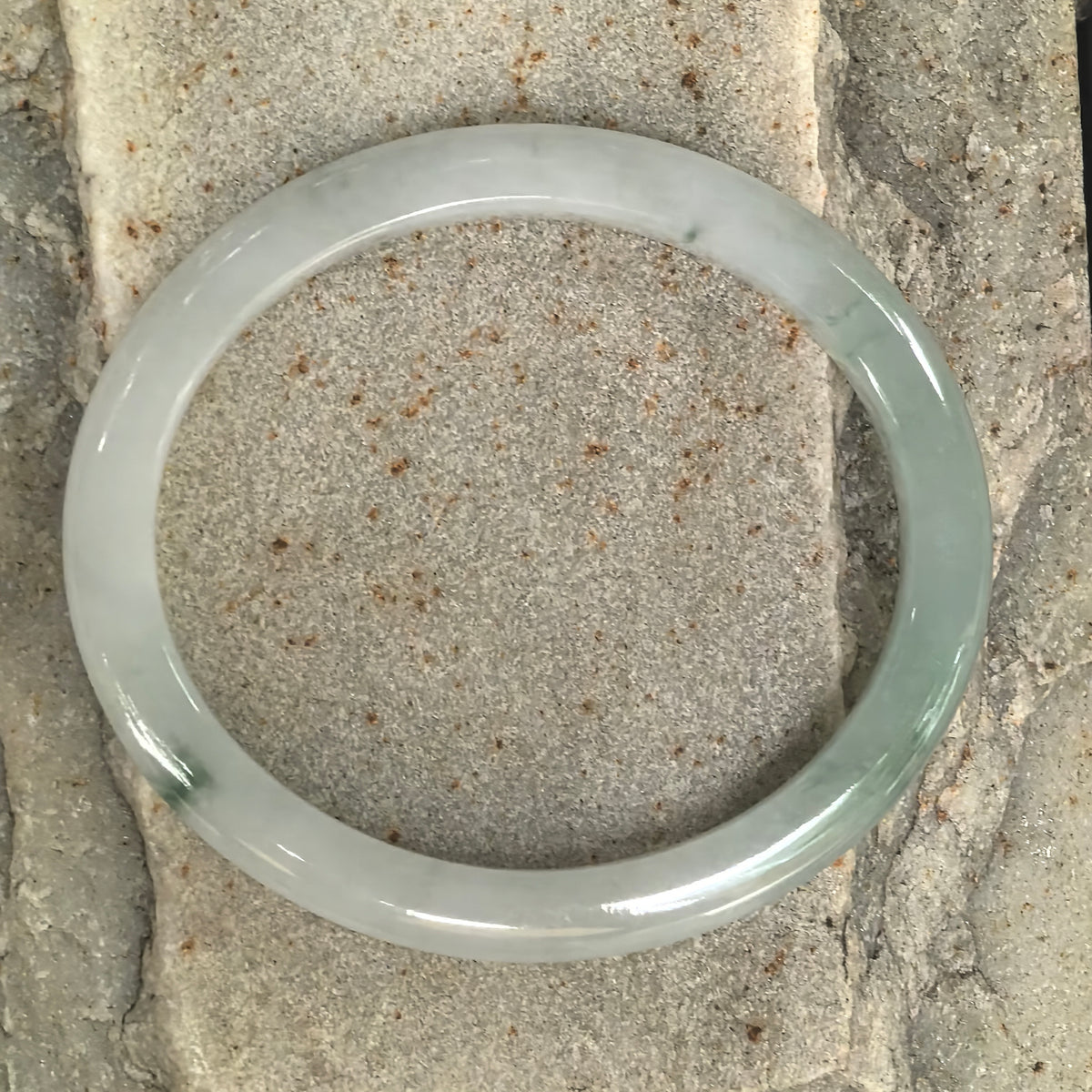 Overhead view of a pale green oval jadeite bangle on textured stone, showing its elongated shape and greener section along one side.