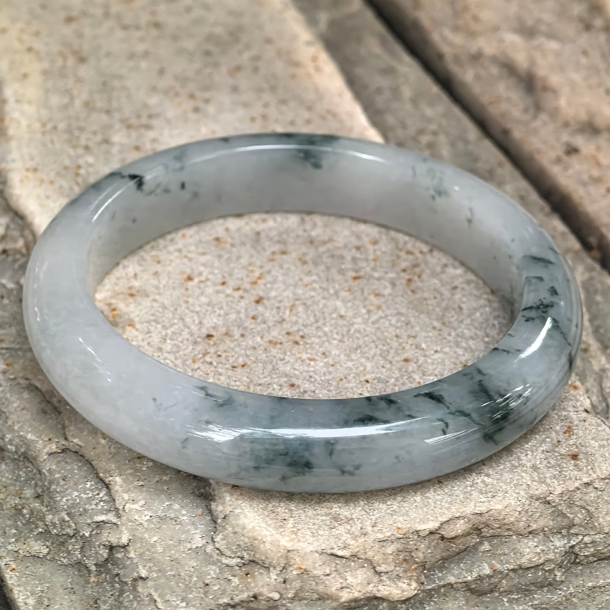 Angled view of the jadeite D-shape bangle on textured stone, showing its rounded exterior and clusters of deep green floating patterns.