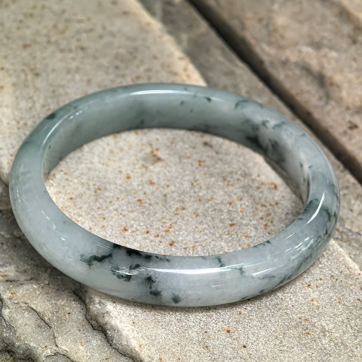 Angled view of the slim jadeite D-shape bangle on textured stone, highlighting its rounded exterior and deep green floating patterns.