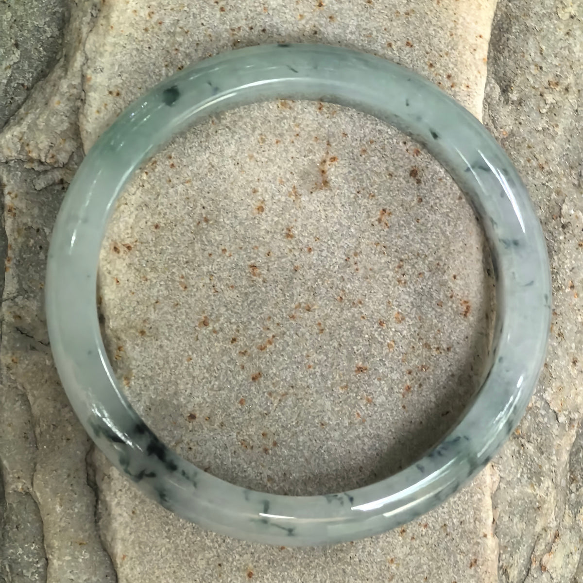 Overhead view of the pale green jadeite bangle on stone, showing its full circular shape and scattered deep green floating patterns.