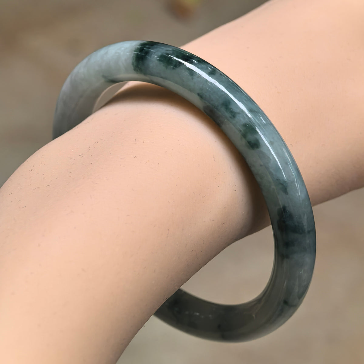 Angled wrist view of a fine glutinous jadeite round-bar bangle with deep green floating patterns over a light green base.
