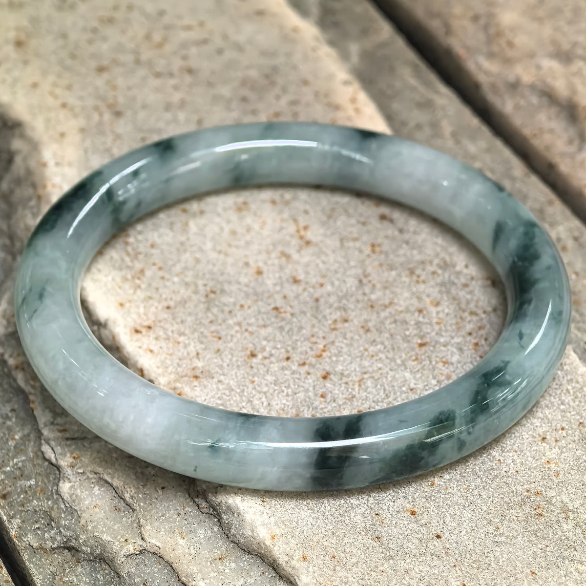 Angled view of the jadeite round-bar bangle on stone, highlighting its rounded profile and contrasting light and deep green tones.