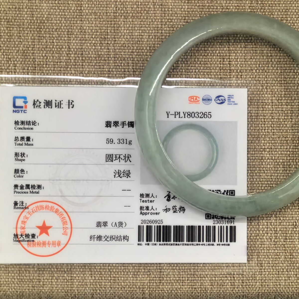 Pale apple green jadeite D-shape bangle displayed over its matching NGTC Grade-A jadeite certificate on a woven background.
