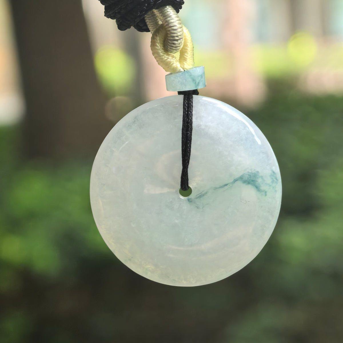 Front view of a glutinous pale green jadeite Peace Pendant hanging against a blurred green background, with a deep green floating pattern extending toward the right edge.