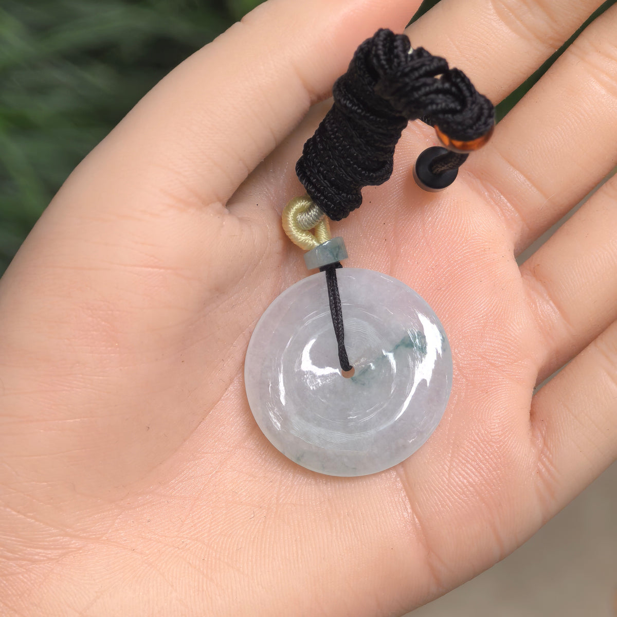 Pale green jadeite Peace Pendant resting in an open palm, showing its deep green floating pattern, black cord, decorative knots and gray-green spacer bead.