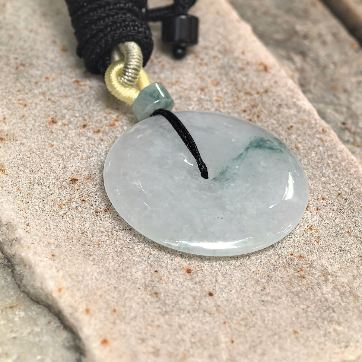 Angled view of a pale green jadeite Peace Pendant on textured stone, showing its rounded edges, polished surface and localized deep green floating pattern.