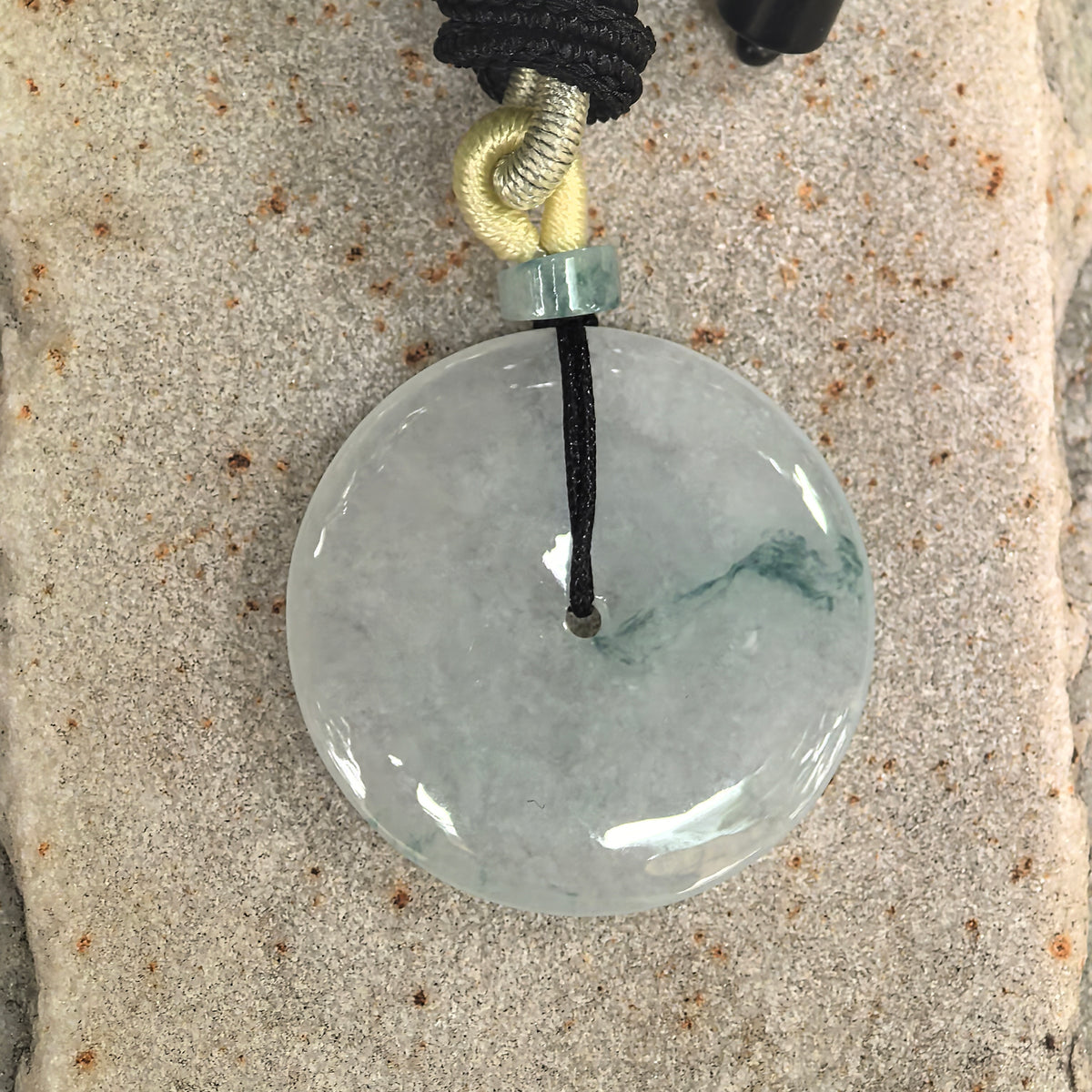 Overhead view of a pale green jadeite Peace Pendant on stone, showing its circular outline, central opening and deep green pattern across the right side.