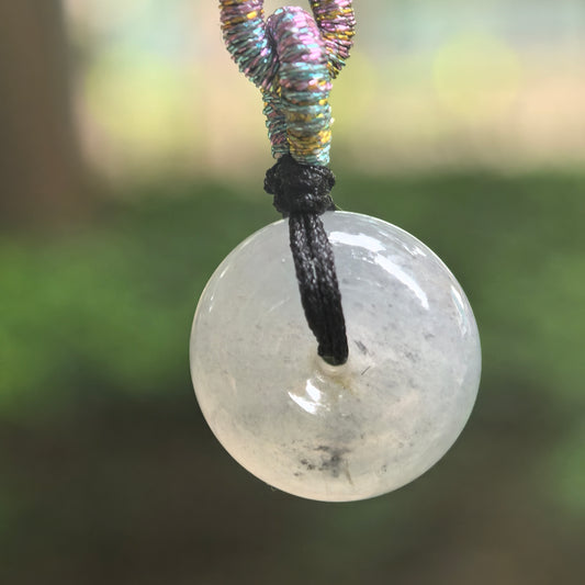 Front view of a small light gray jadeite Peace Pendant hanging from a black cord, showing an ink-black floating pattern against a blurred green background.